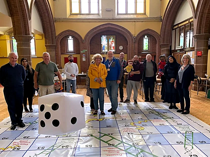 A hall with an oversized snakes-and-ladders board spread across the floor and a giant soft dice on it, with a group gathered behind the game.